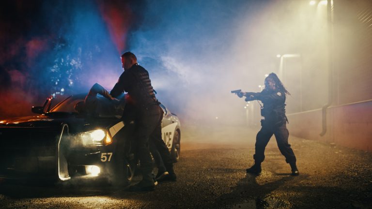 Pessoa sendo algemada por policial, enquanto outra aponta uma arma