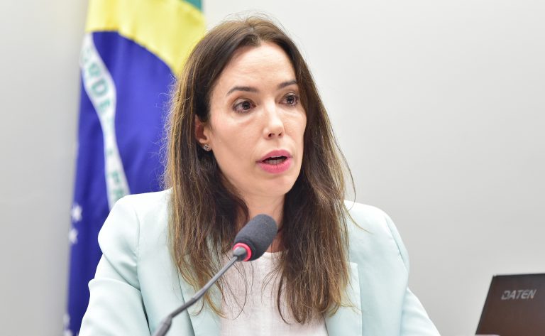 Uma mulher fala ao microfone. Ela é branca, tem cabelos comprido e castanho e usa um blazer azul claro