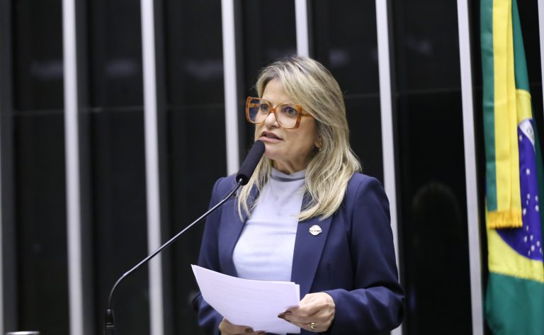 Deputada Flávia Morais fala ao microfone. Ela é loira, tem o cabelo comprido, usa uma blusa branca e segura um papel