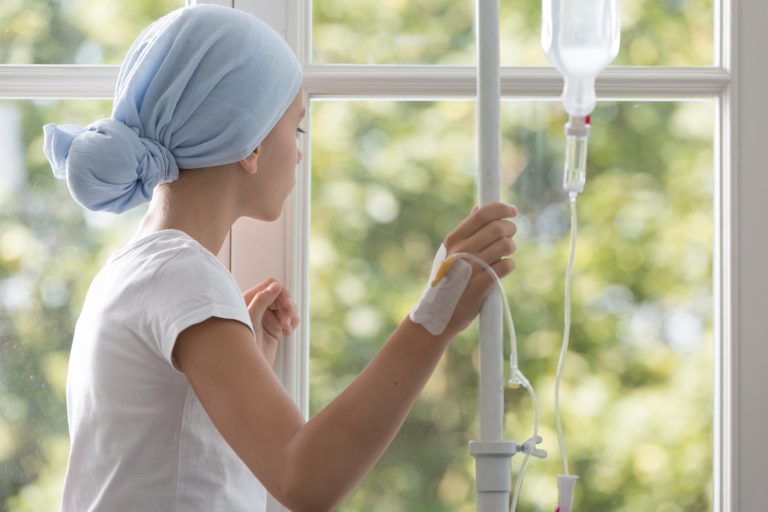 Menina com lenço na cabeça e segurando medicação venosa olha pela janela