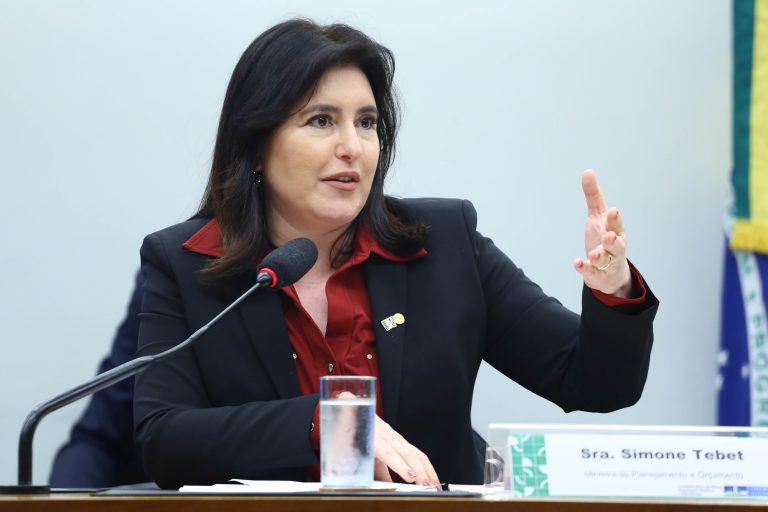 Ministra do Planejamento, Simone Tebet. Ela é uma mulher clara, com cabelos longos, lisos e escuros. Usa um blazer preto e uma camisa vermelha