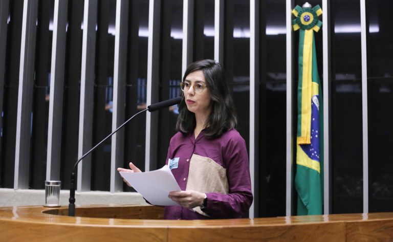 Uma mulher branca discursa. Ela tem cabelo escuro e veste uma camisa rosa