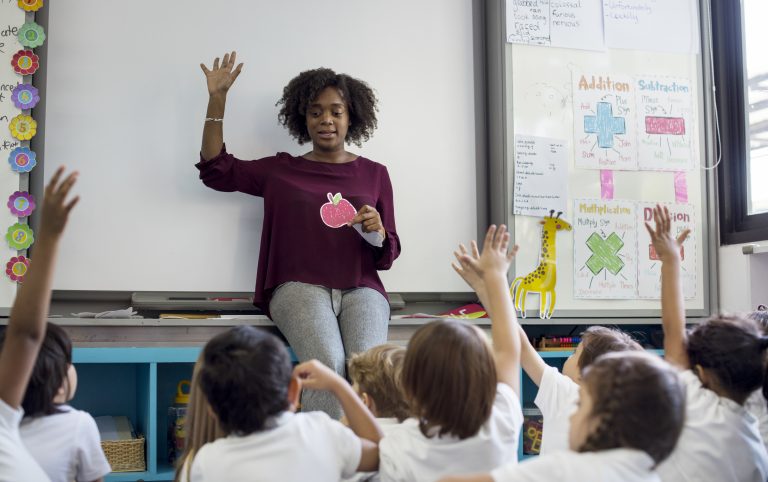 Professora está de costas para o quadro e de frente para os alunos. Ela segura o desenho de uma maça e está com o outro braço levantado. Algumas crianças também estão com os braços levantados