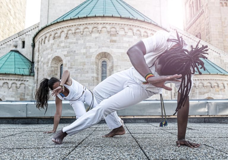Um homem e uma mulher negros lutam capoeira. Eles vestem roupas brancas e estão abaixados com uma das mãos no chão e a outra acima da cabeça