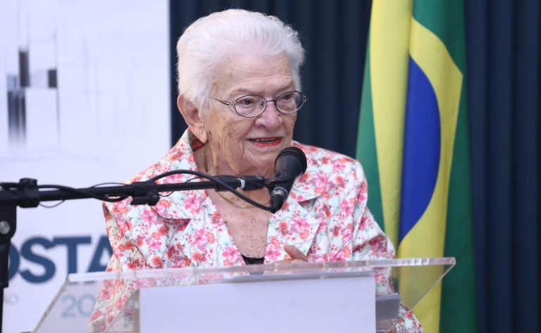 Deputada Luiza Erundina fala ao microfone. Ela é uma mulher idosa, clara e com cabelos brancos. Ela usa óculos e um blazer com flores cor-de-rosa. Atrás dela, há uma bandeira do Brasil num mastro