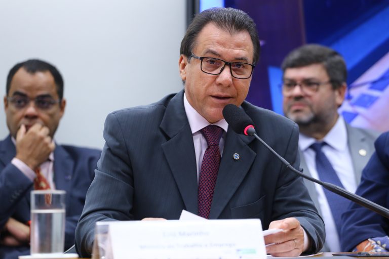 Ministro do Trabalho, Luiz Marinho, fala ao microfone. Ele é um homem claro, com cabelo escuro, usa um terno escuro, camisa rosa claro e gravata vinho. Ele usa óculos e segura um papel.