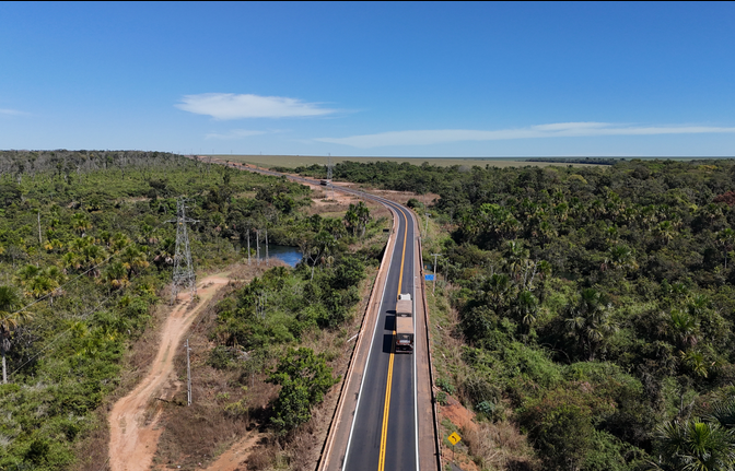 Imagem aérea de uma rodovia de pista simples atravessando uma área de vegetação nativa.Caminhões e outros veículos seguem pela estrada asfaltada, com faixa amarela central.Ao redor, há mata densa, estrada de terra, postes de energia e céu azul com poucas nuvens.